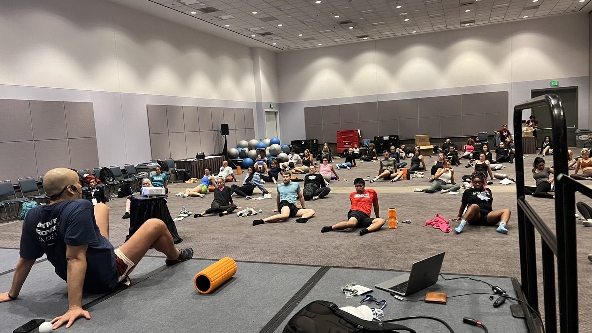 Marc Coronel seated on the floor with a foam roller, leading a room of fitness professionals through an Active Recovery workshop